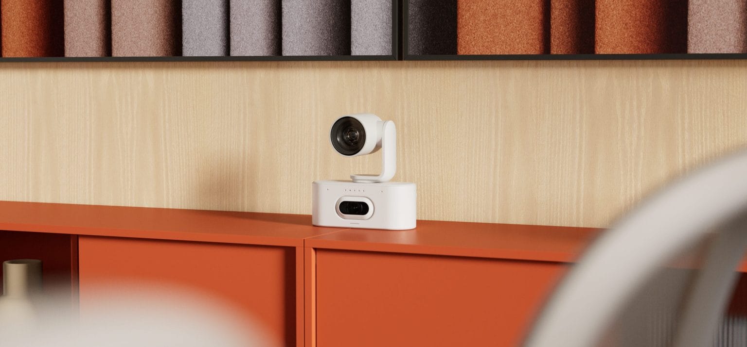 logitech rally ai camera - מצלמת וידאו קונפרנס לבנה מונחת על מזנון כתום בחדר ישיבות מעוצב עם חיפוי אקוסטי ברקע