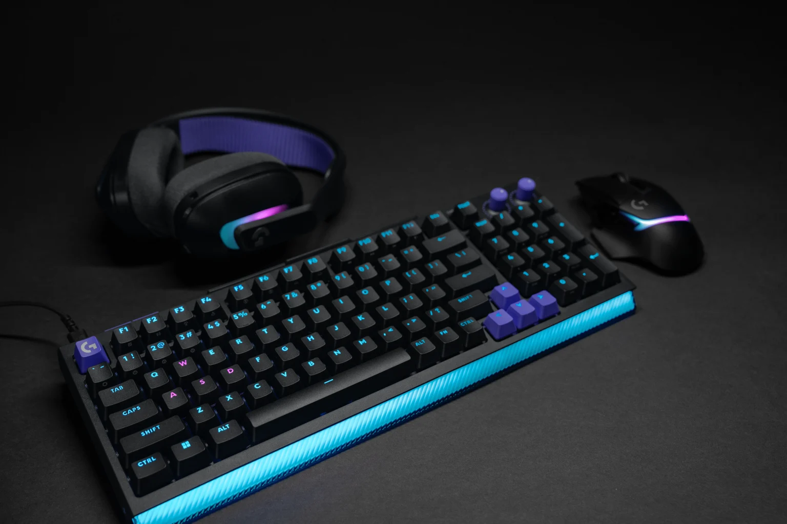 Logitech G512 X - מקלדת גיימינג שחורה בפריסת 98% עם מקשים סגולים, תאורת RGB כחולה, ולצדה אוזניות ועכבר תואמים על רקע שחור.