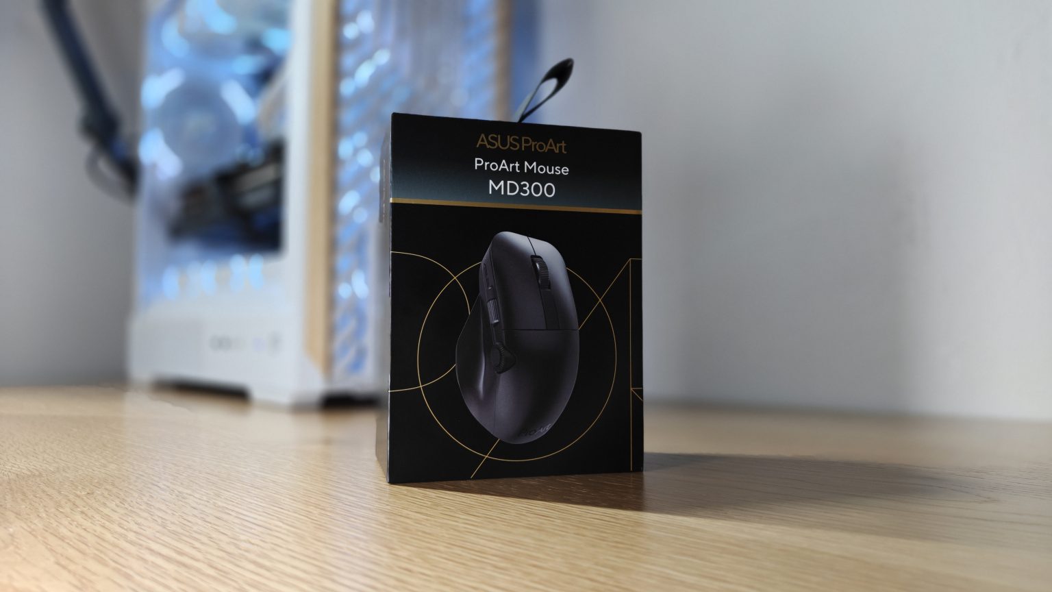 Asus ProArt Mouse MD300 - אריזת העכבר מונחת על שולחן עץ בסביבת עבודה, עם מחשב נייח ברקע