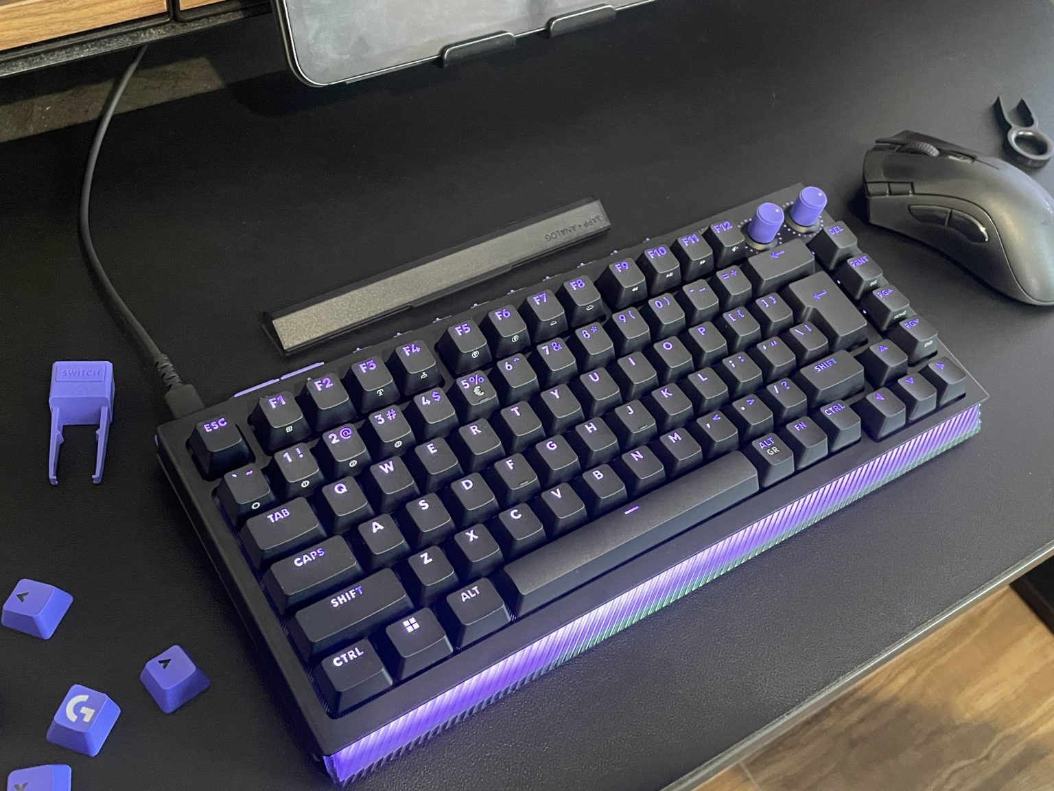 Logitech G512X - מקלדת גיימינג מכנית שחורה עם מקשים סגולים ותאורת צד, מונחת על משטח עבודה לצד עכבר גיימינג וחולץ מקשים
