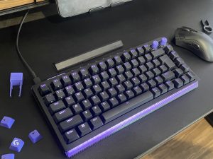 Logitech G512X - מקלדת גיימינג מכנית שחורה עם מקשים סגולים ותאורת צד, מונחת על משטח עבודה לצד עכבר גיימינג וחולץ מקשים
