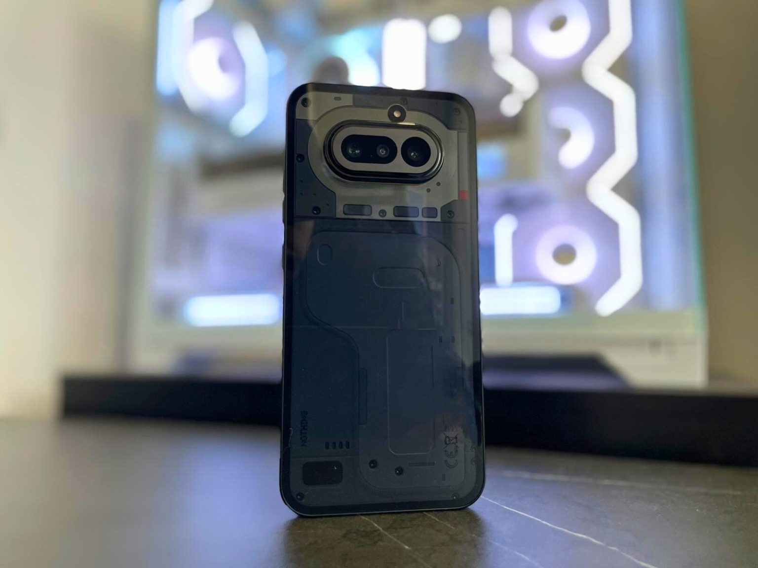 Nothing Phone 4a - גב המכשיר עם העיצוב השקוף, מערך המצלמות הכפול והרכיבים הפנימיים החשופים, מונח על שולחן.