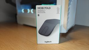 Logitech Mobi Fold - קופסת אריזה של עכבר נייד מתקפל אפור, מונחת על שולחן עץ עם מחשב גיימינג מואר ברקע