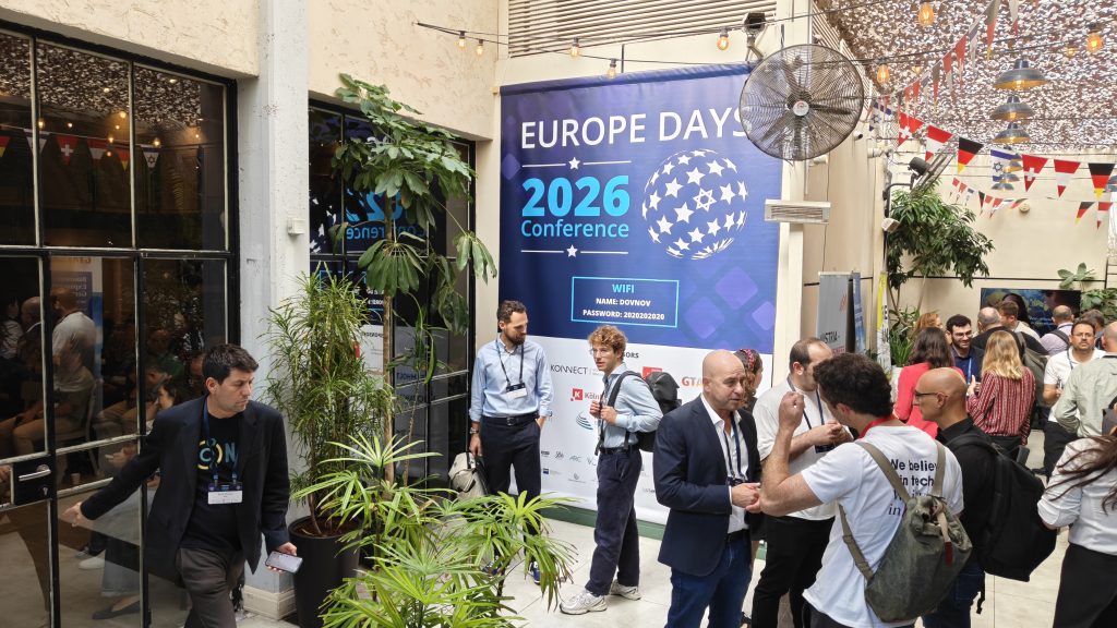 EuropeDays 2026 - קהל משתתפים, אורחים ואנשי עסקים מתמנגלים ומשוחחים בחצר האירוע, מול שלט כחול גדול של כנס Europe Days 2026 Conference עם סמלי נותני חסות, צמחייה עשירה ודגלי מדינות תלויים.